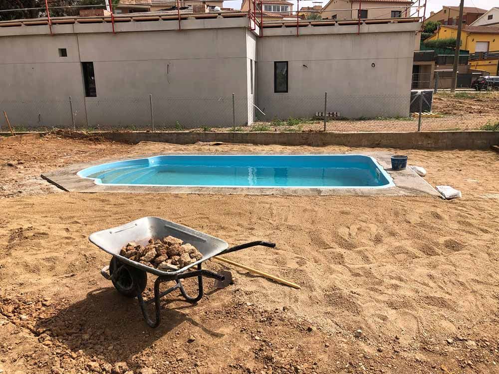 Una carretilla llena de piedras en un terreno baldío junto a una pequeña poza azul y un edificio blanco.