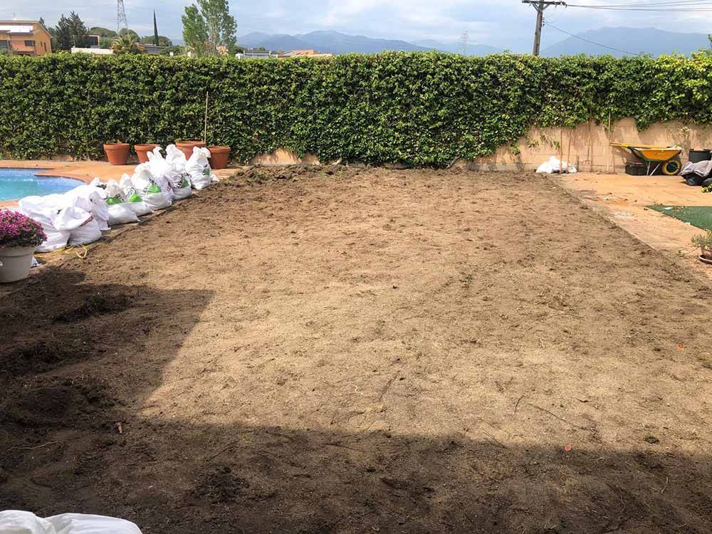 Terreno de tierra delimitado por setos, con sacos apilados a la izquierda y una zona sombreada delante.