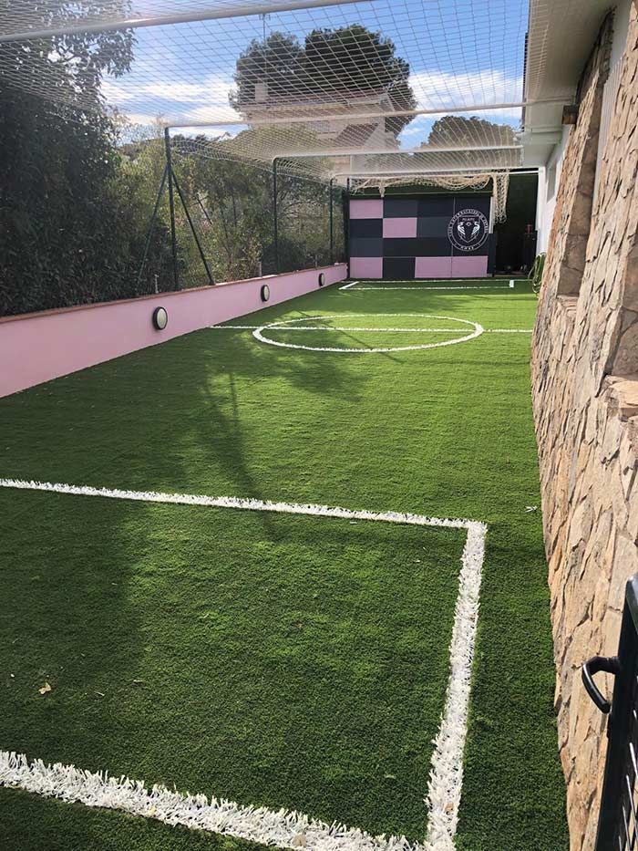 Pequeña cancha de fútbol en el patio trasero con césped artificial, líneas blancas, una red que la protege y una pared rosa y negra al final.
