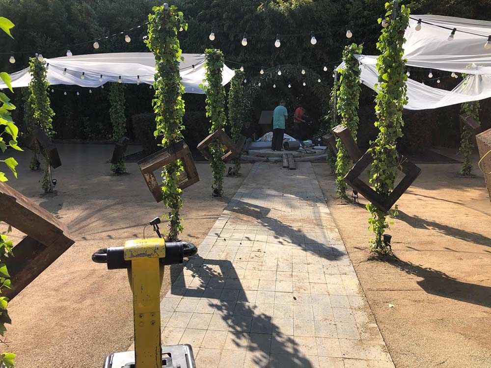 Patio exterior con postes cubiertos de hiedra, guirnaldas de luces y una persona de pie cerca de unas mesas bajo un toldo blanco.