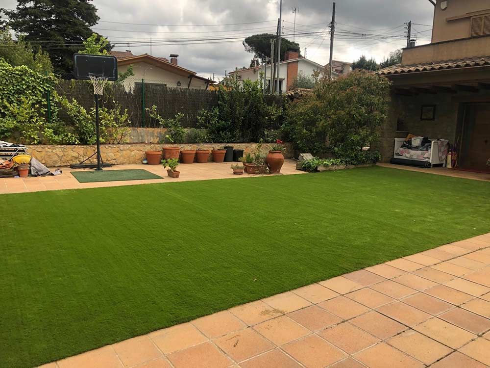 Patio trasero con césped verde brillante, adoquines de terracota, plantas en macetas y un patio cubierto bajo un cielo nublado.