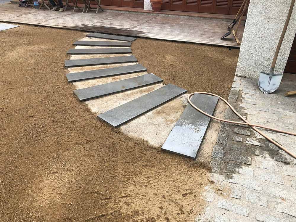 Hilera curva de losas grises dispuestas sobre tierra marrón junto a un edificio blanco y una manguera.