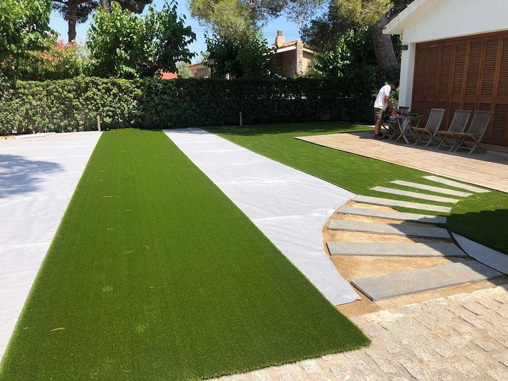 Patio trasero con franjas de césped artificial verde, bordes blancos, patio y una persona cerca de la casa.