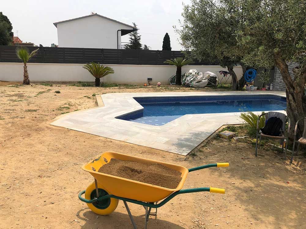 Patio trasero con piscina, jardín de tierra y una carretilla amarilla en primer plano.