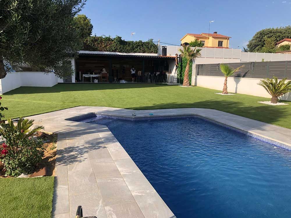 Piscina en el patio trasero con agua azul, terraza de baldosas, césped y un patio sombreado bajo un cielo despejado.