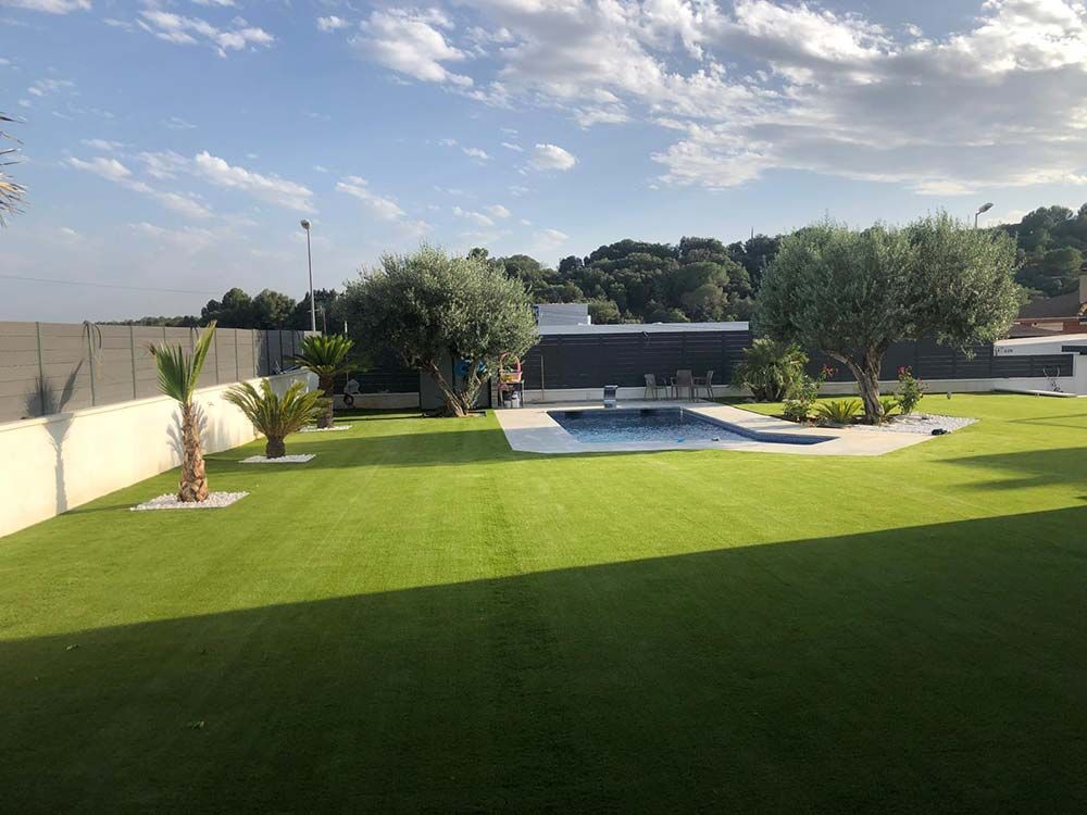 Jardín trasero moderno con un gran césped verde, piscina, patio y árboles bajo un cielo parcialmente nublado.