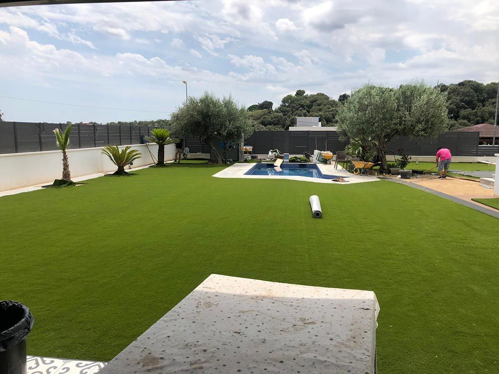 Patio trasero con césped artificial, piscina, tumbonas y vallas blancas bajo un cielo nublado.