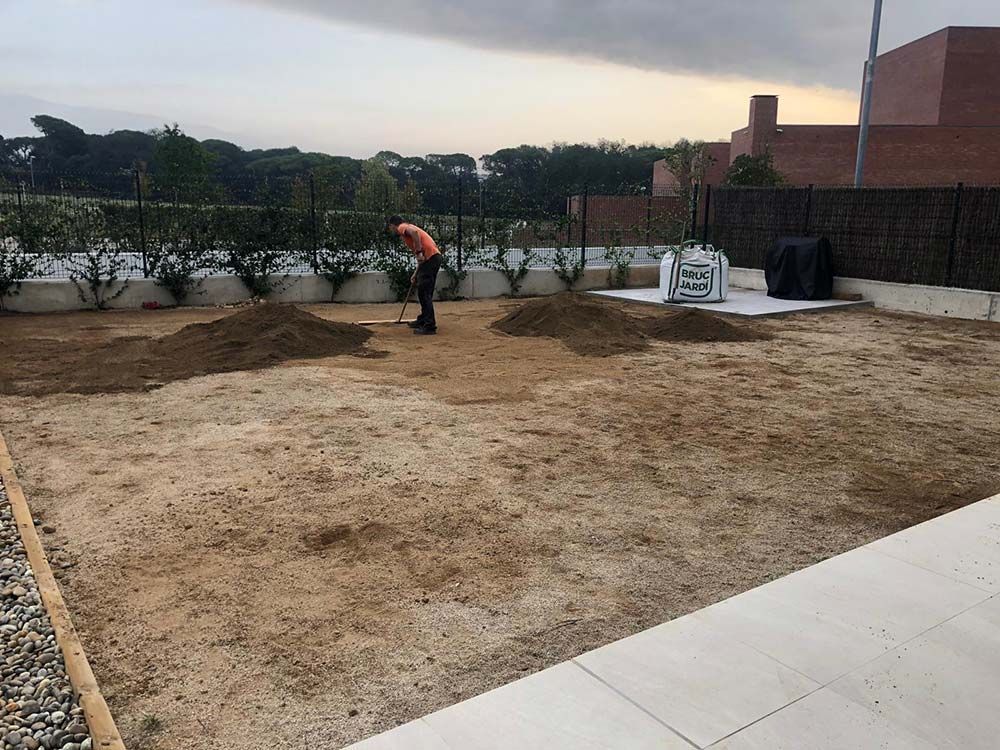 Un trabajador esparce tierra en un patio trasero en construcción, cerca de una cerca y edificios de ladrillo.