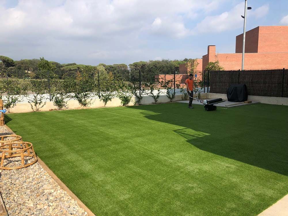 Jardín trasero con césped artificial, setos y construcciones de ladrillo bajo un cielo parcialmente nublado.