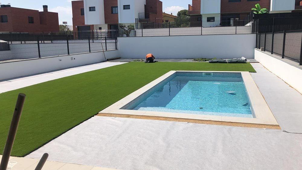 Piscina en el patio trasero junto a césped artificial y un patio blanco, con edificios de apartamentos al fondo.