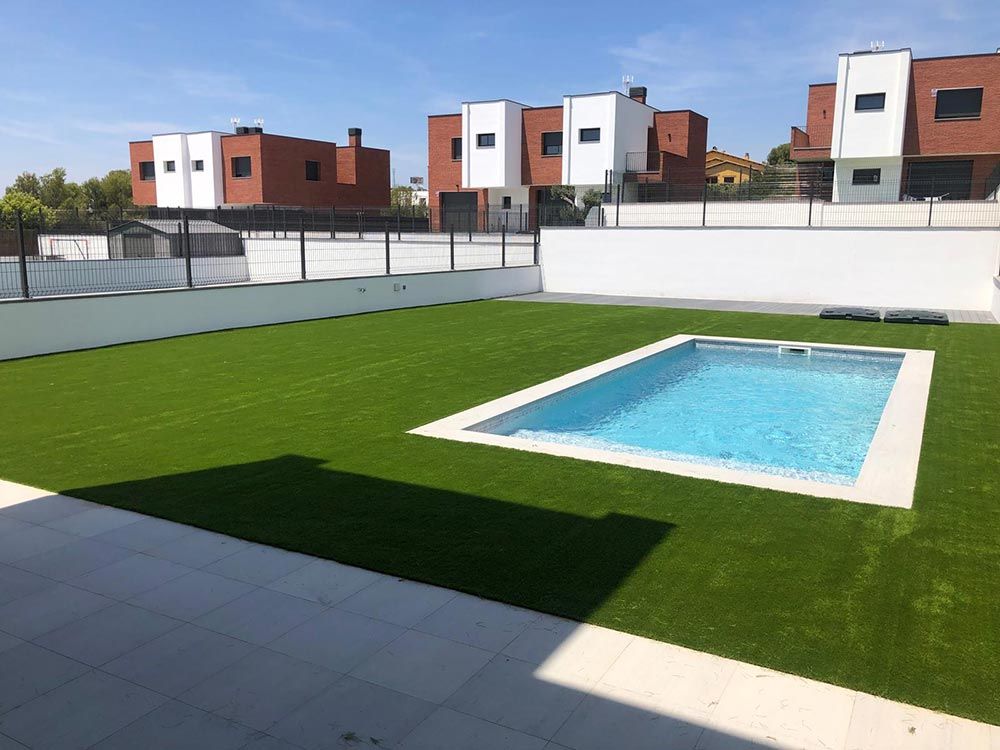 Patio con césped verde y una pequeña piscina azul, rodeado de muros blancos y edificios modernos.