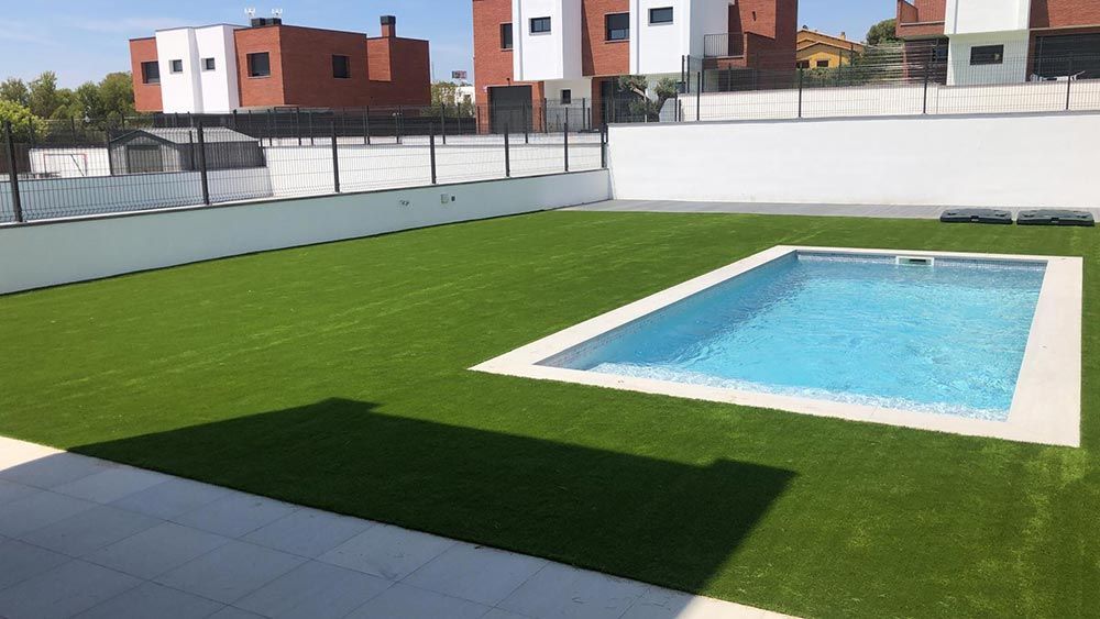 Patio trasero con piscina rectangular, césped verde y valla blanca bajo un sol brillante.
