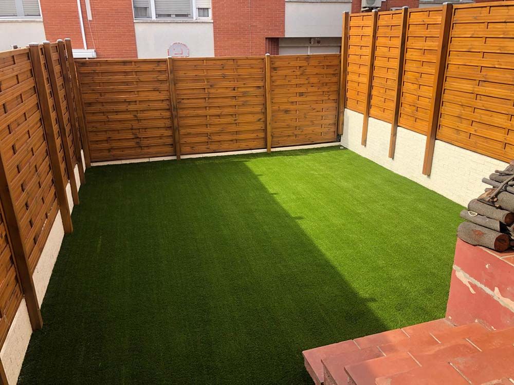 Patio trasero con césped artificial, valla de madera y un borde pavimentado soleado.