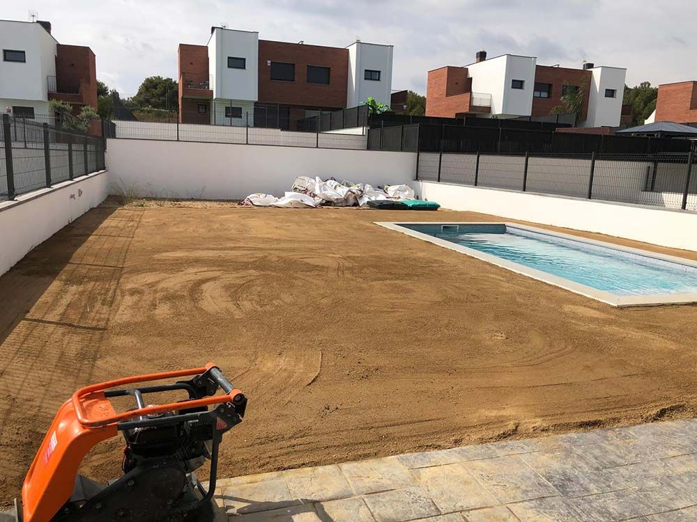 El patio trasero está siendo nivelado para construir una piscina, con una pequeña piscina y una excavadora naranja en primer plano.