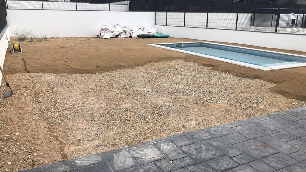 Patio trasero con grava y tierra, una pequeña piscina rectangular y un borde de patio pavimentado.