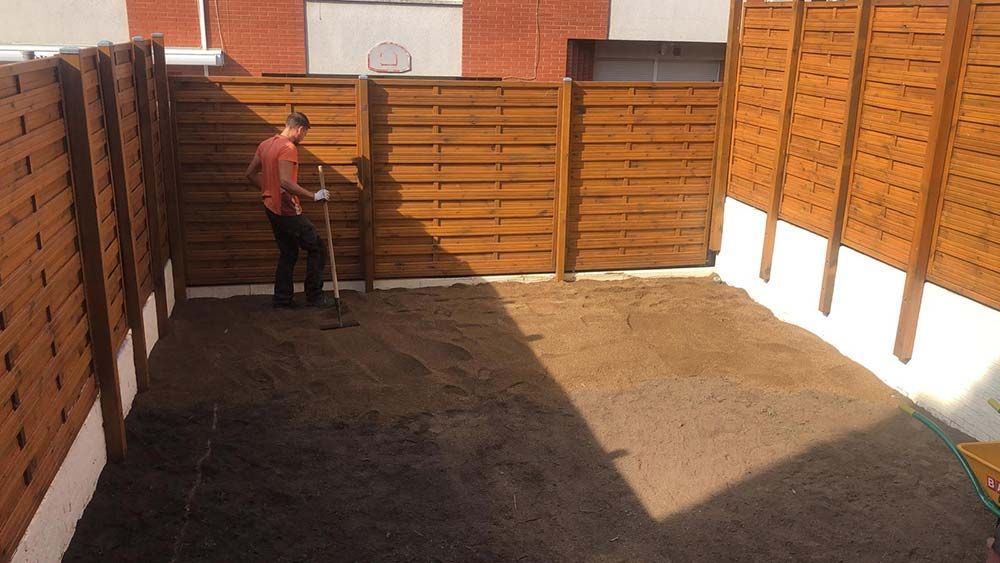 Una persona se encuentra en un patio trasero soleado, de pie junto a una puerta y una cerca de madera, sosteniendo una herramienta.