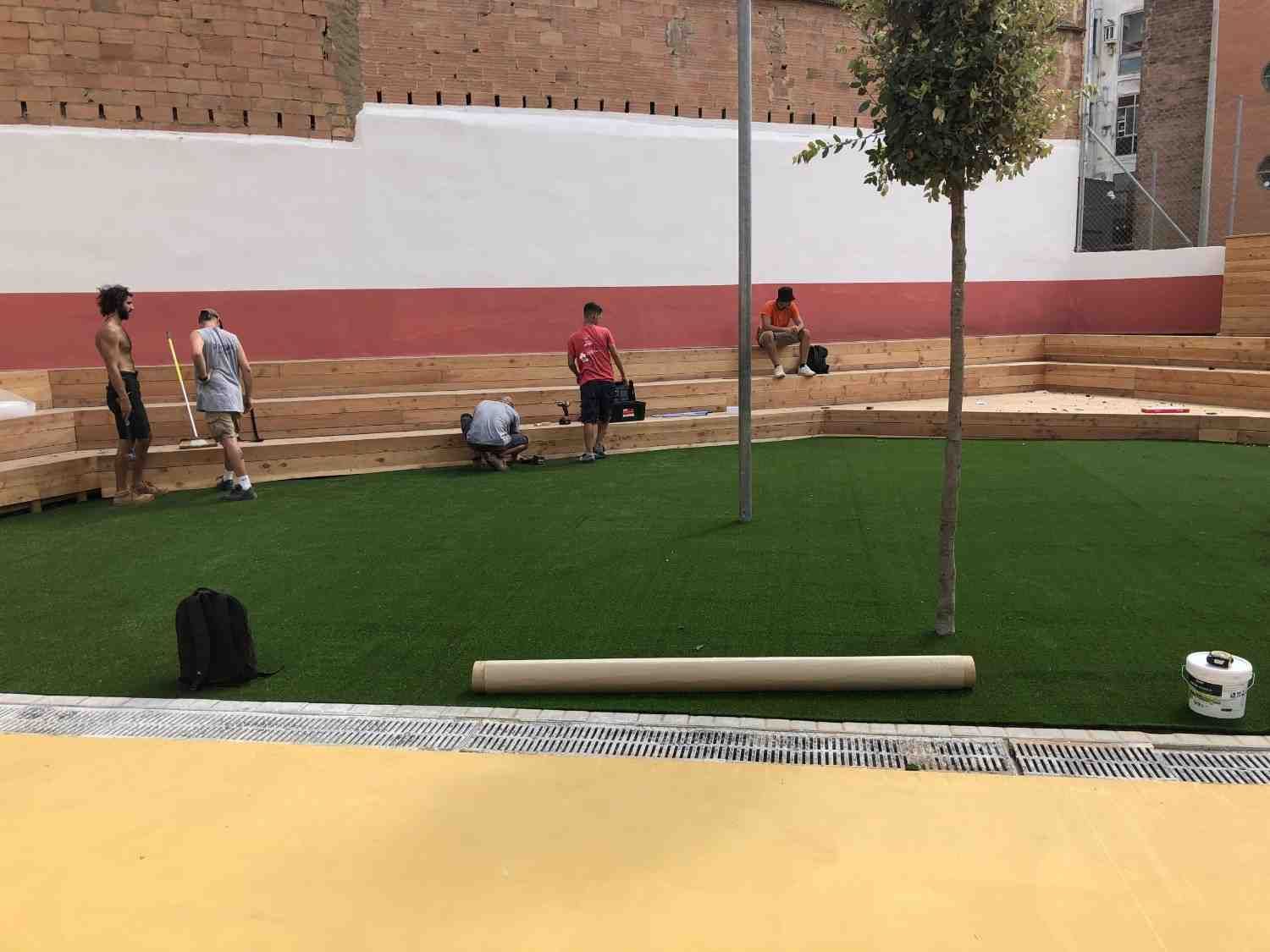 Gente en un patio ajardinado con césped verde, un sendero amarillo y bancos junto a una pared de ladrillos.