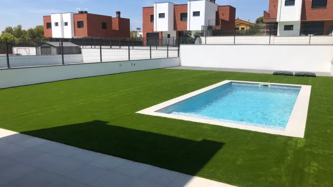Piscina exterior con agua azul, terraza blanca y césped artificial verde en un complejo residencial.