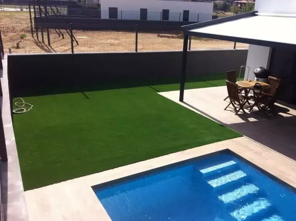 Patio trasero con césped artificial verde junto a una piscina azul y un conjunto de comedor de patio sombreado.