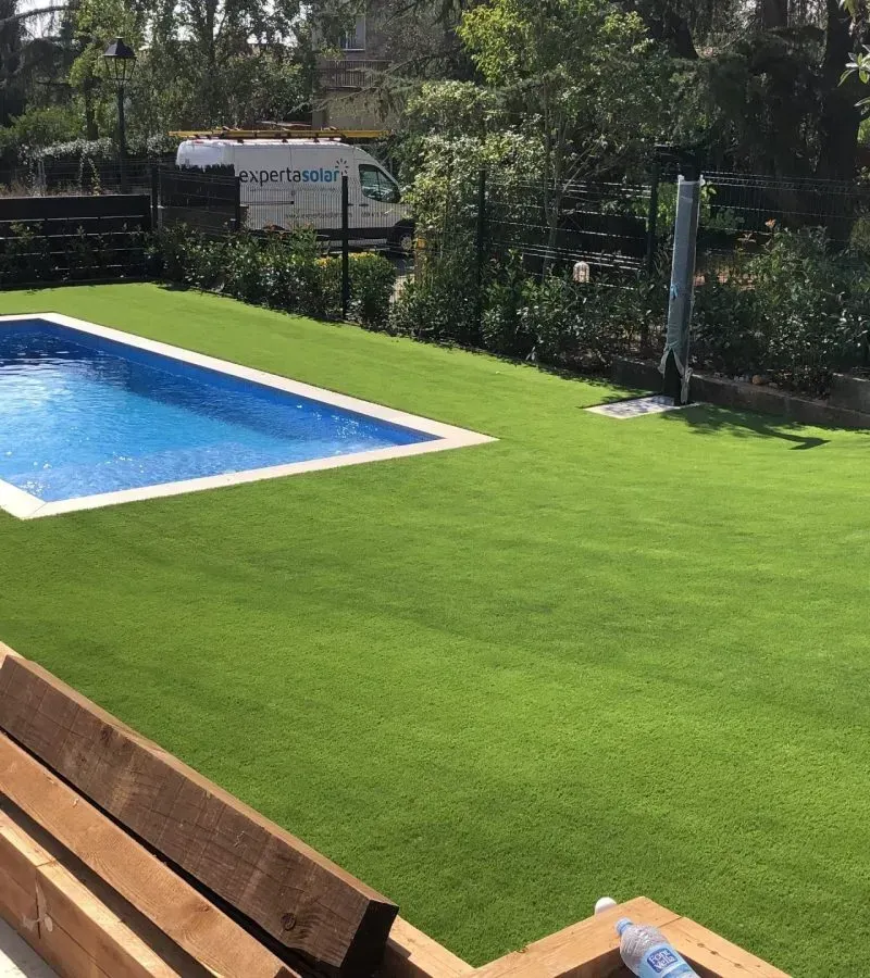 Piscina en el patio trasero junto a césped artificial de color verde brillante y banco de madera bajo los árboles.