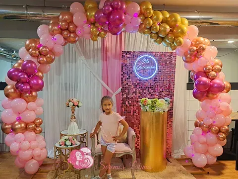 Una niña está sentada en una silla bajo un arco de globos rosas y dorados.