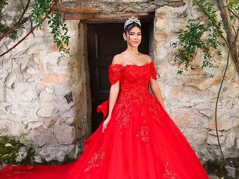 Una mujer con un vestido de fiesta rojo y una tiara está parada frente a un muro de piedra.
