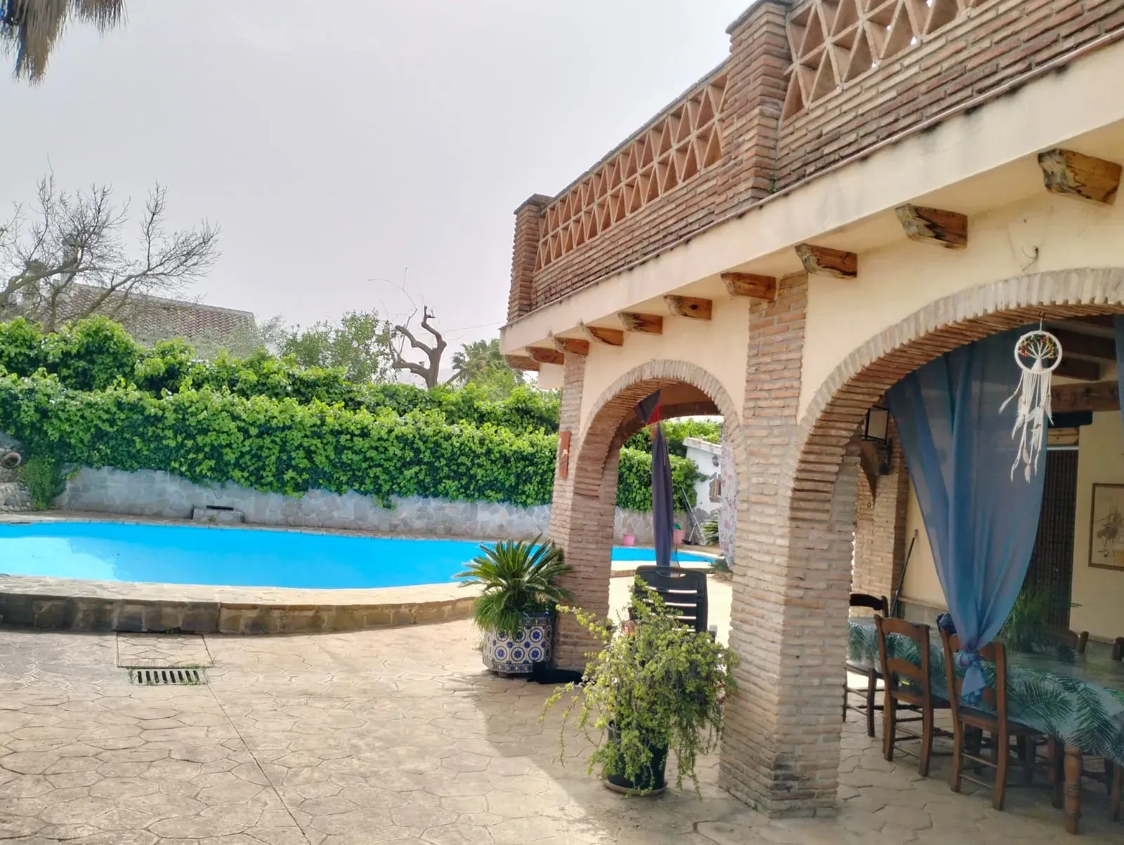 Edificio junto a la piscina con arcos de ladrillo, piscina, cortina azul y exuberante vegetación.
