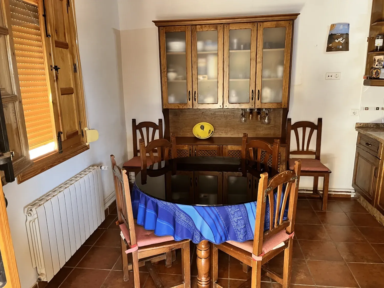 Un comedor con una mesa redonda negra, sillas de madera y un armario. La luz del sol entra por una ventana.