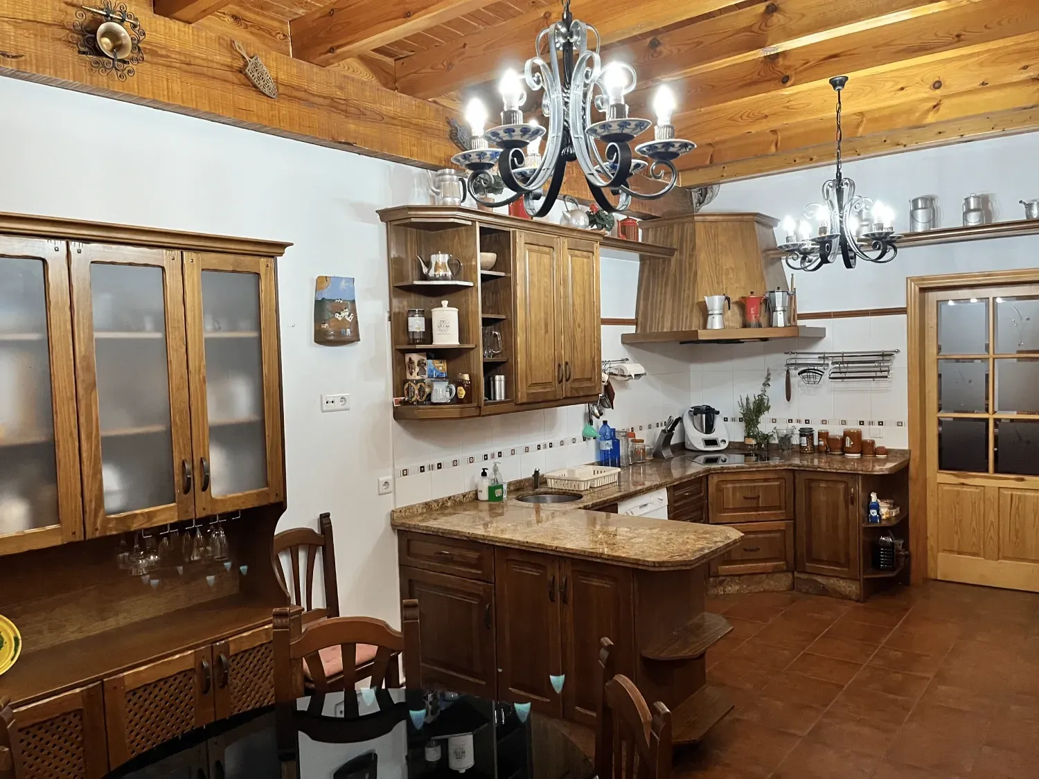 Cocina rústica con gabinetes de madera, isla y techo con vigas. Dos lámparas de araña cuelgan.