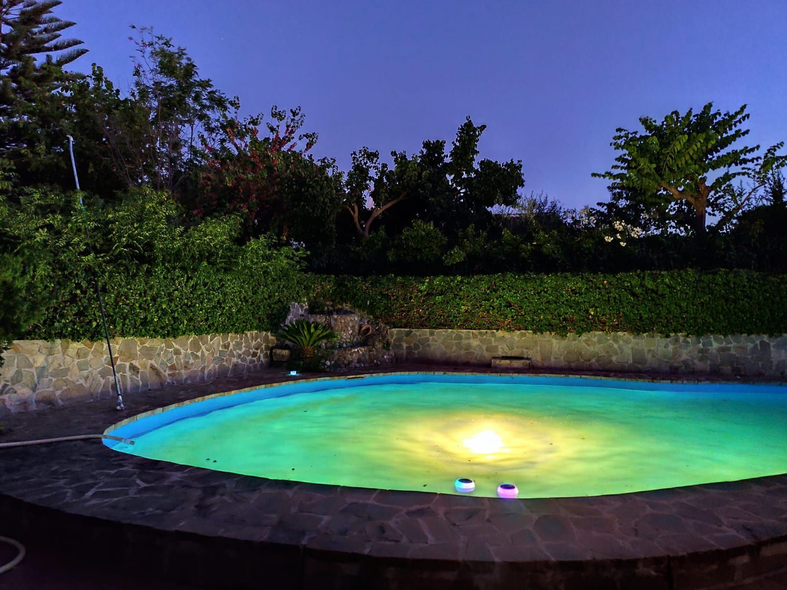 Piscina redonda iluminada por la noche, rodeada de piedra, exuberante vegetación y un cielo oscuro.