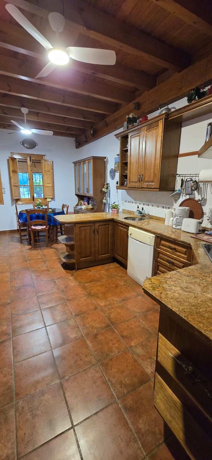 Una cocina con gabinetes de madera, piso de baldosas, ventilador de techo y una mesa de comedor al fondo.