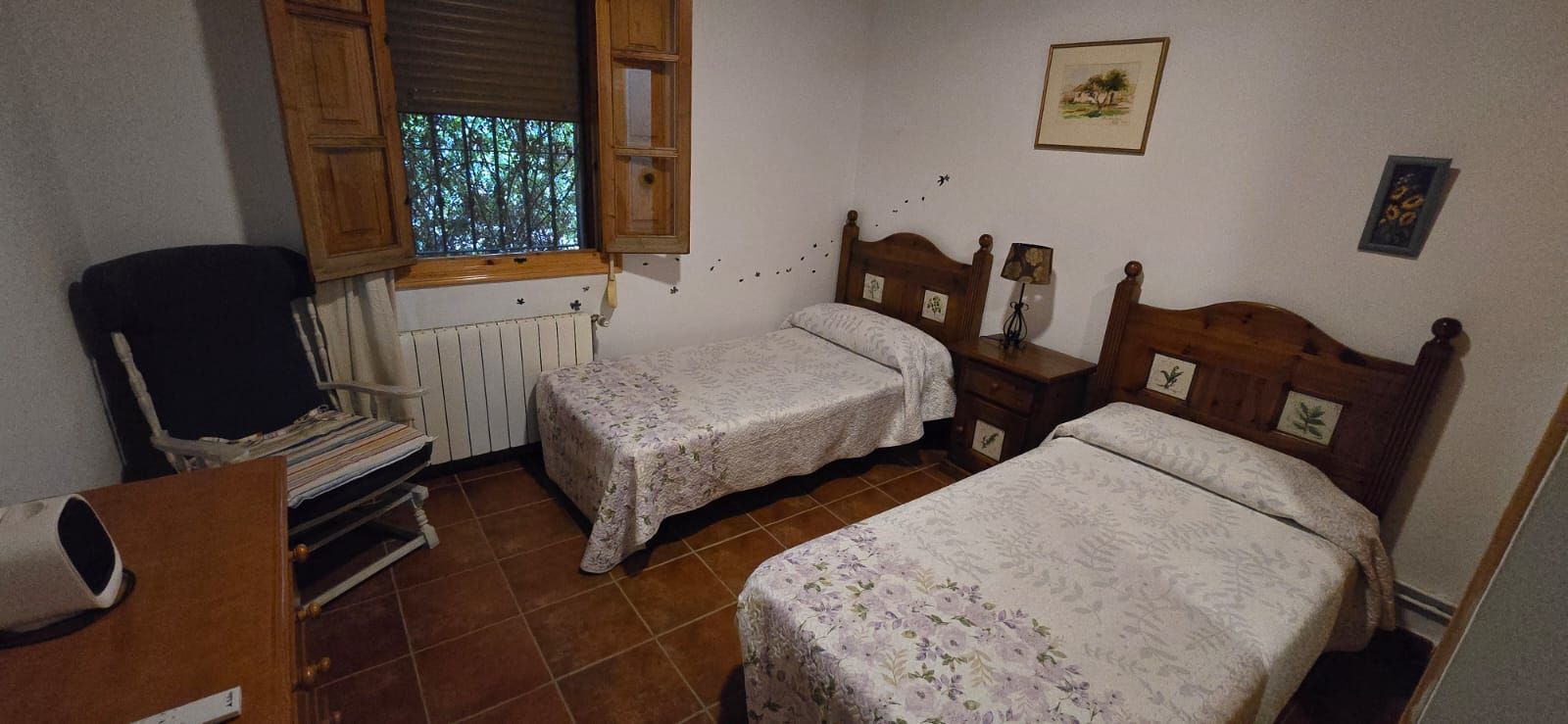 Dormitorio con dos camas individuales, mesita de noche con lámpara, ventana con persianas y silla.