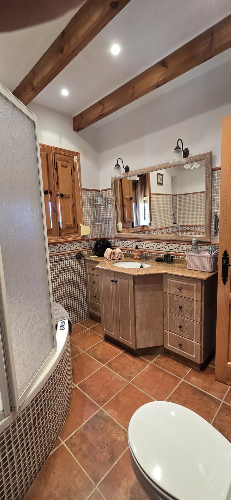 Un baño con vigas de madera a la vista, suelo de baldosas y tocador con espejo.