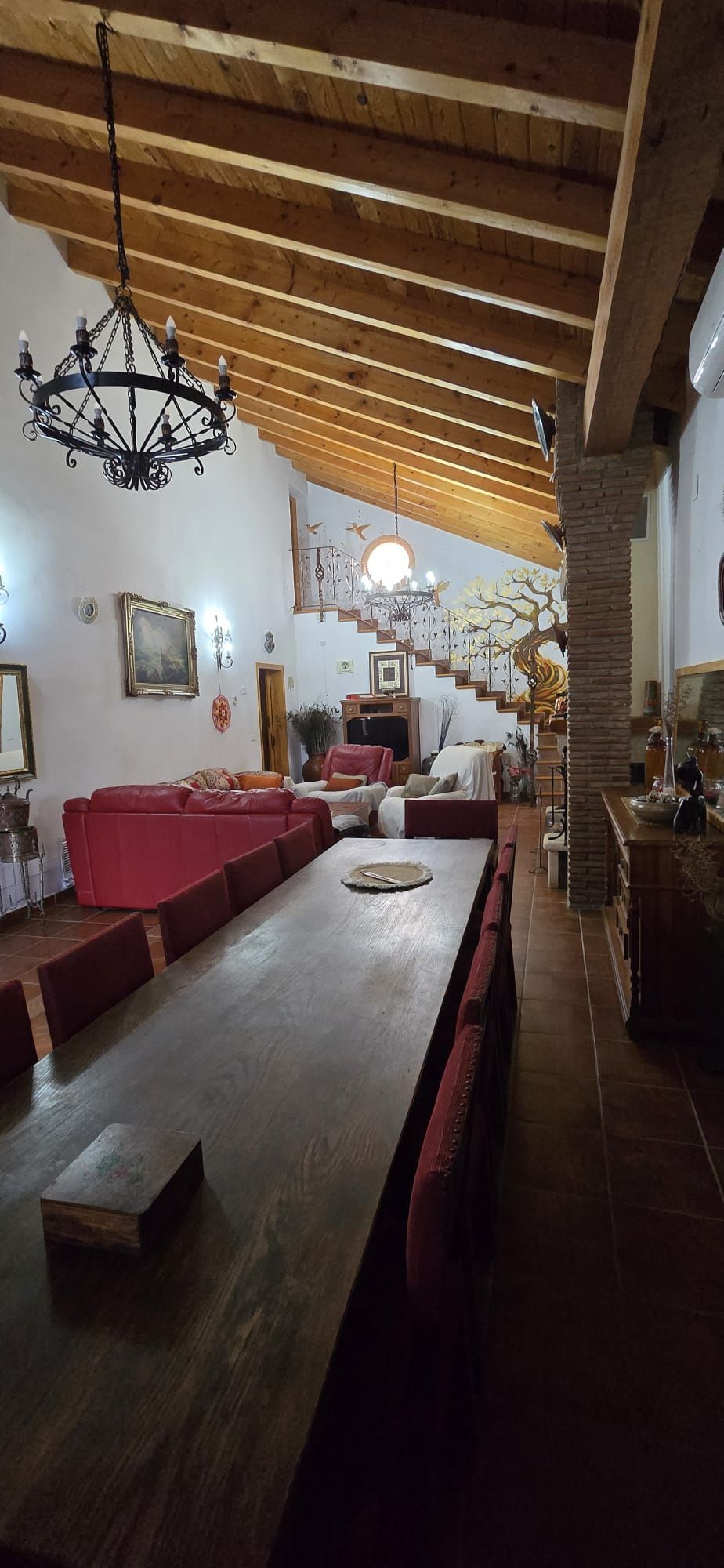 Vista interior de un comedor con una gran mesa de madera, sillas rojas y una escalera.