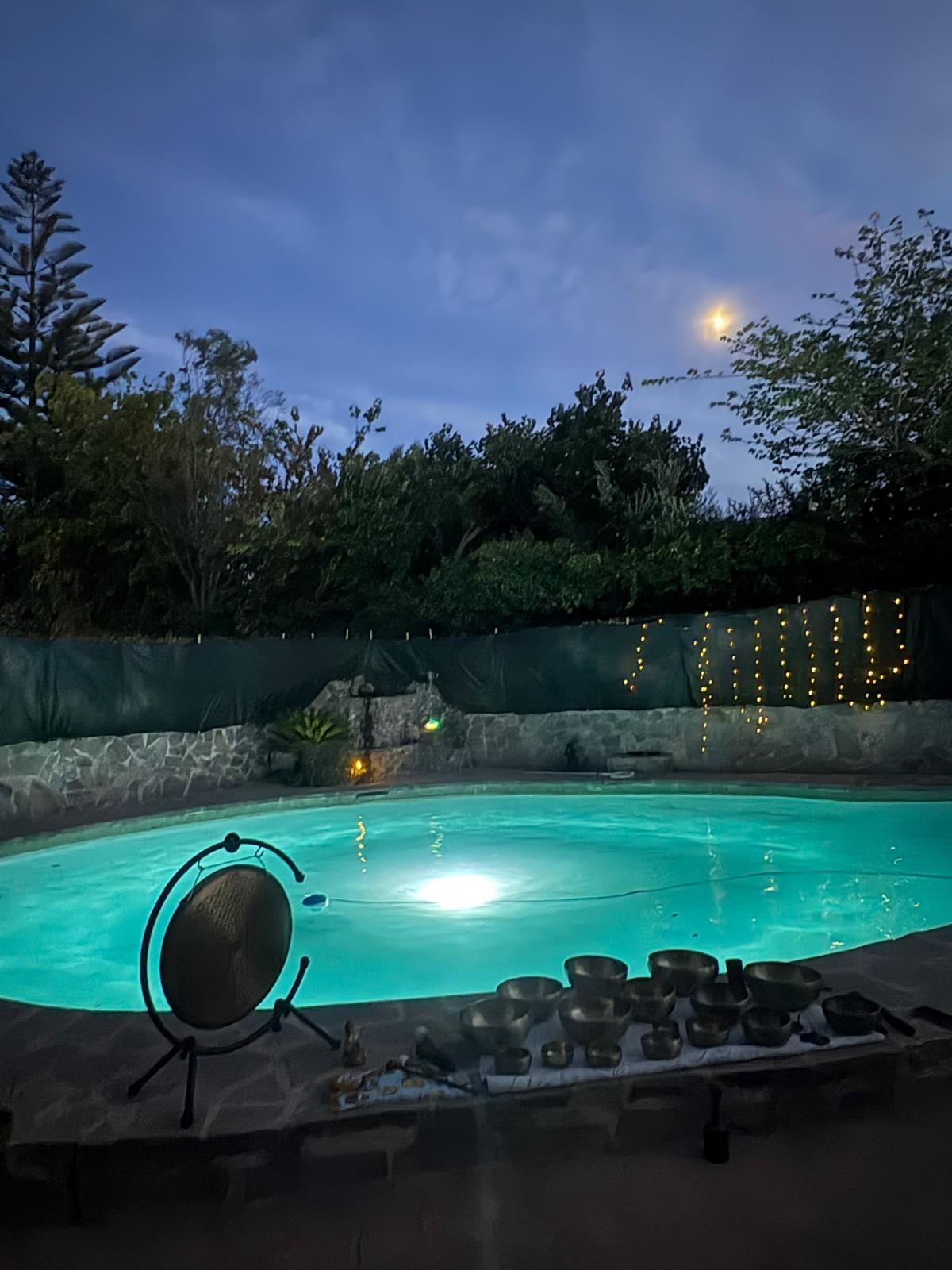 Piscina iluminada por la noche con un gong, cuencos sonoros, luces de cadena y árboles bajo un cielo iluminado por la luna.