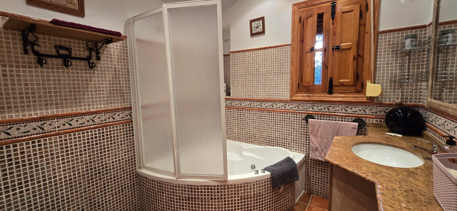 Baño con bañera de esquina, lavabo y ventana con contraventanas de madera, paredes alicatadas.