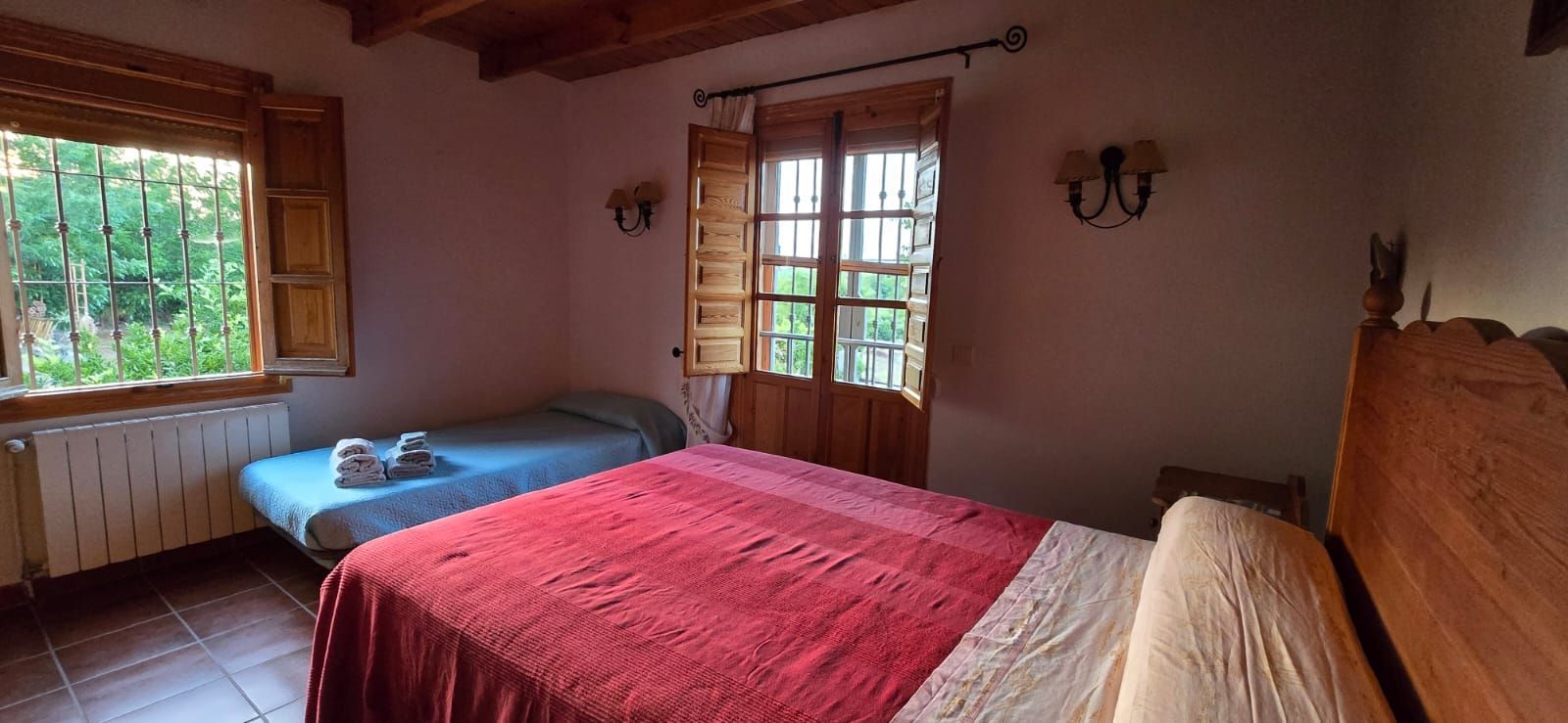 Dormitorio con dos camas, una de matrimonio con funda roja y otra individual, ventana y apliques de pared.