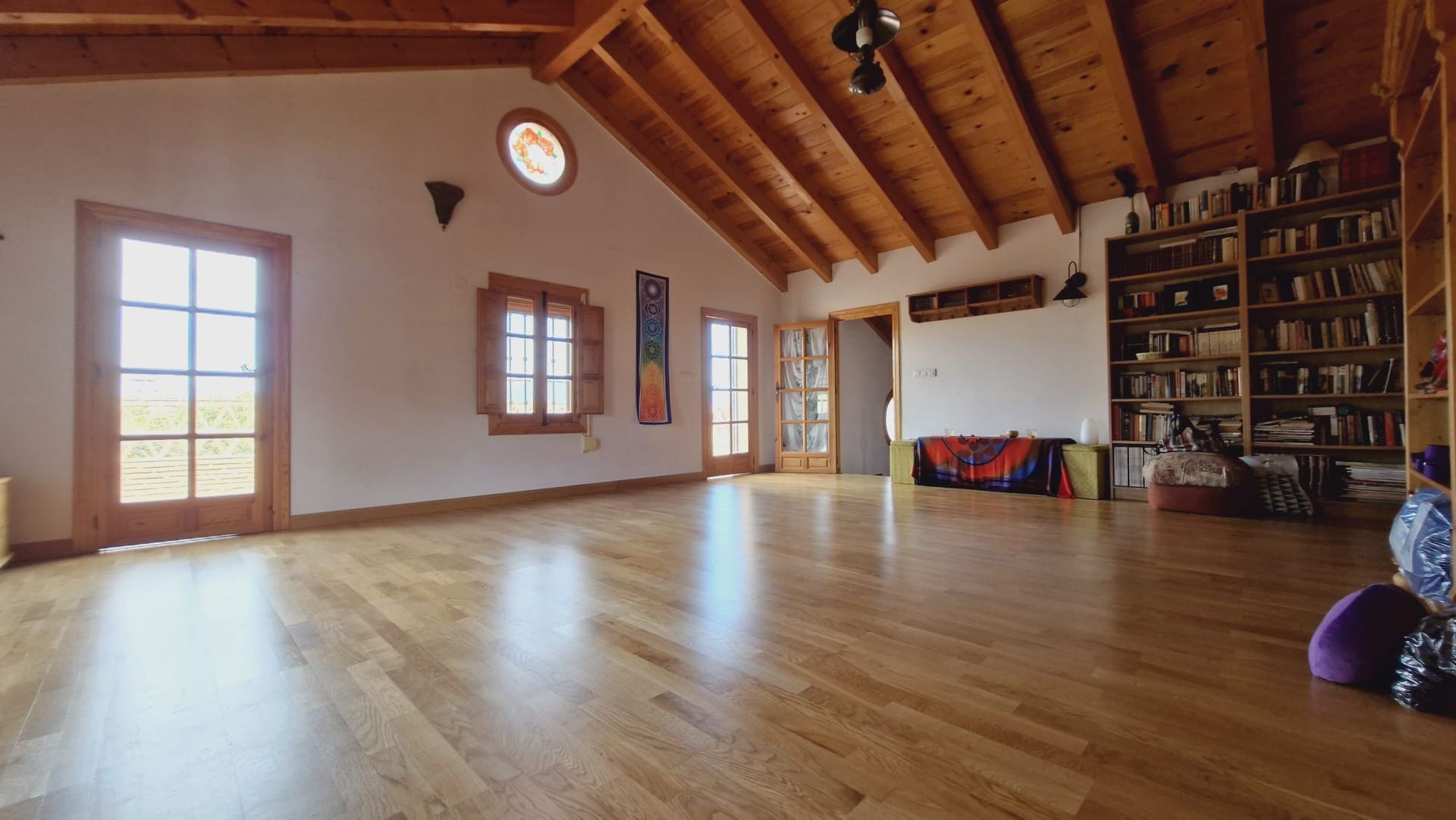 Habitación espaciosa con suelos de madera, techos altos de madera y estanterías. Grandes ventanales con vistas; dos puertas.