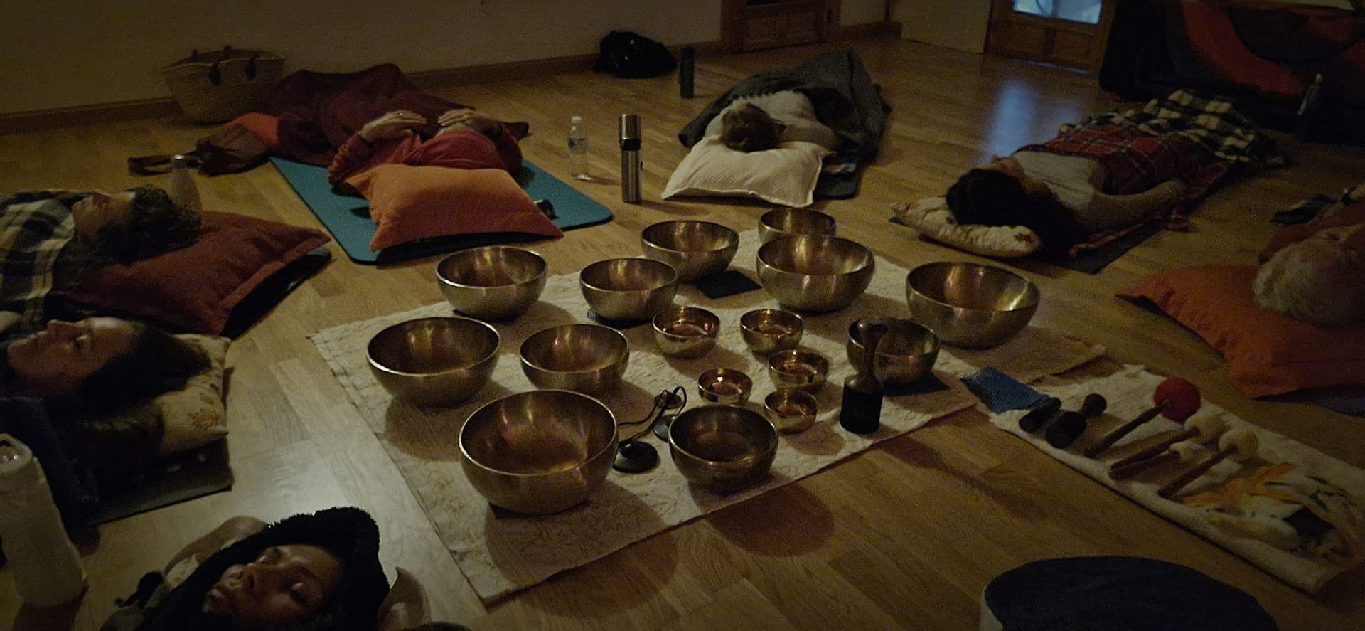 Personas tumbadas en el suelo rodeadas de cuencos cantores, en un entorno de meditación o sanación sonora.