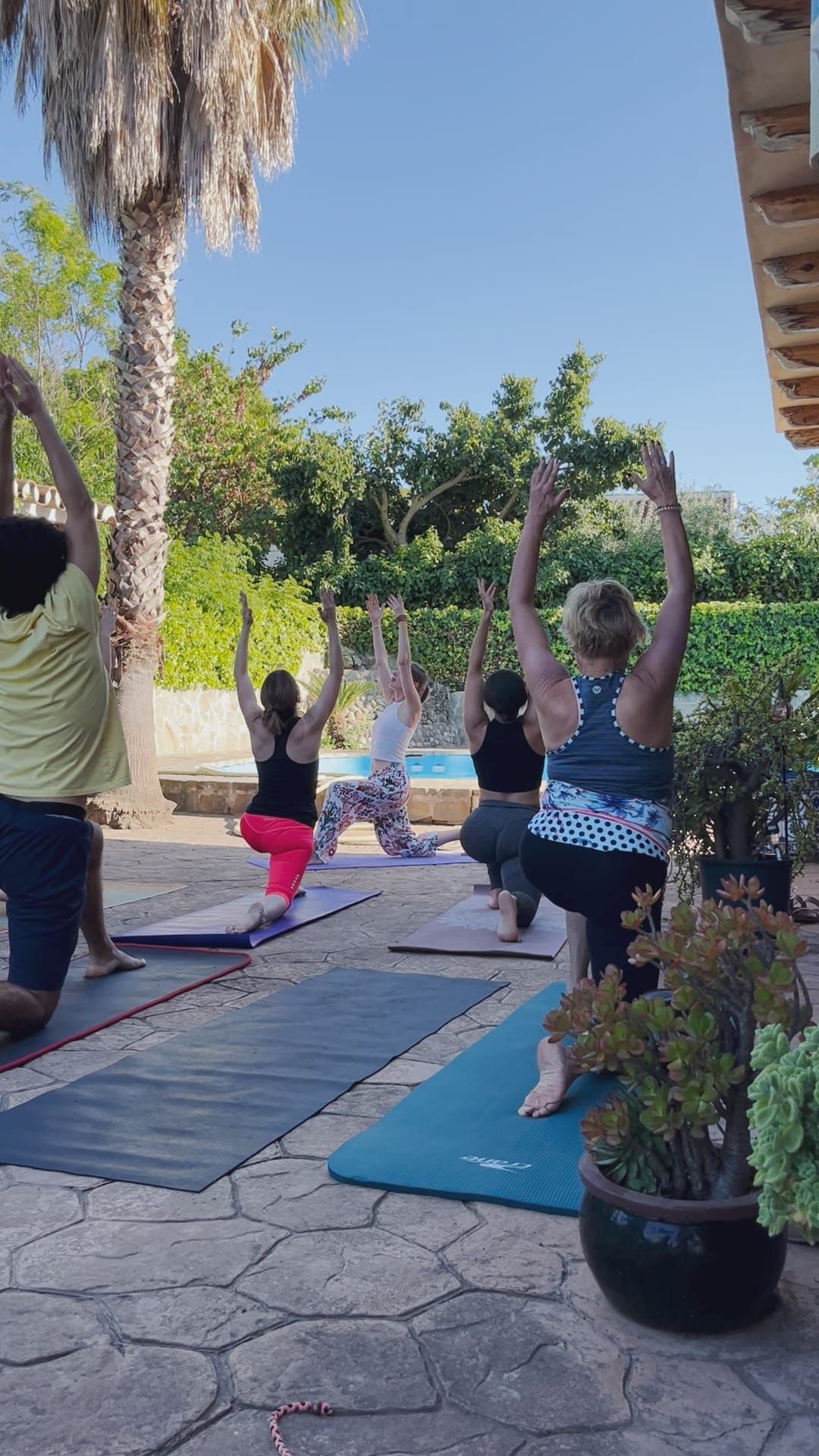 Personas practicando yoga al aire libre, con los brazos en alto, cerca de una piscina. Día soleado.