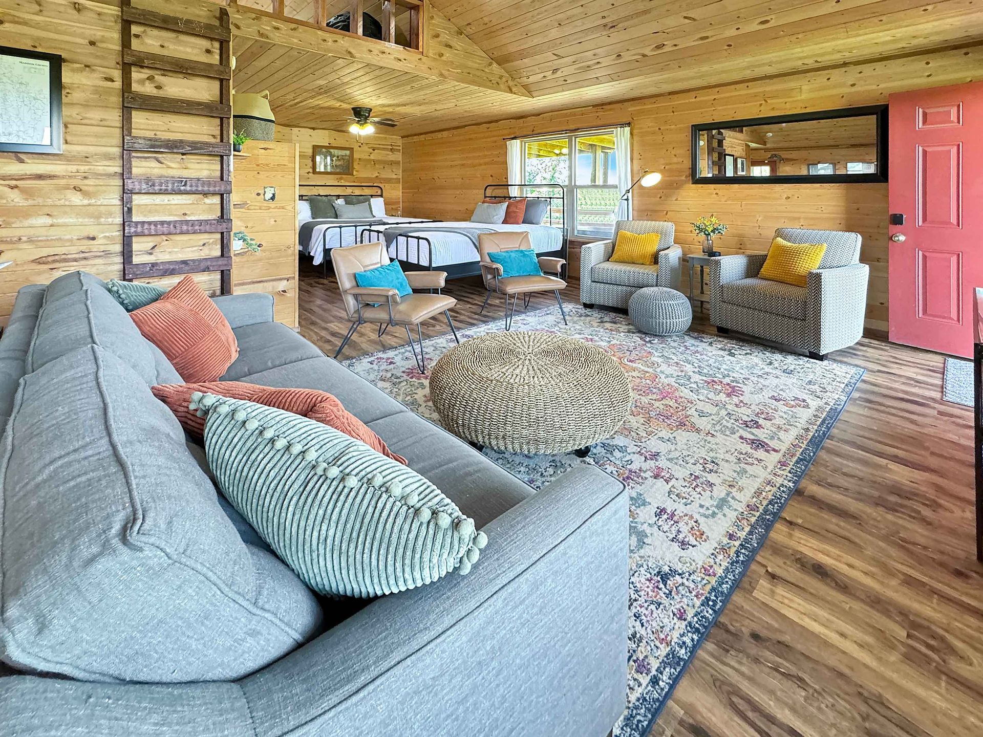 Cozy cabin living room with a gray couch, rug, armchairs, and a bed in the background.
