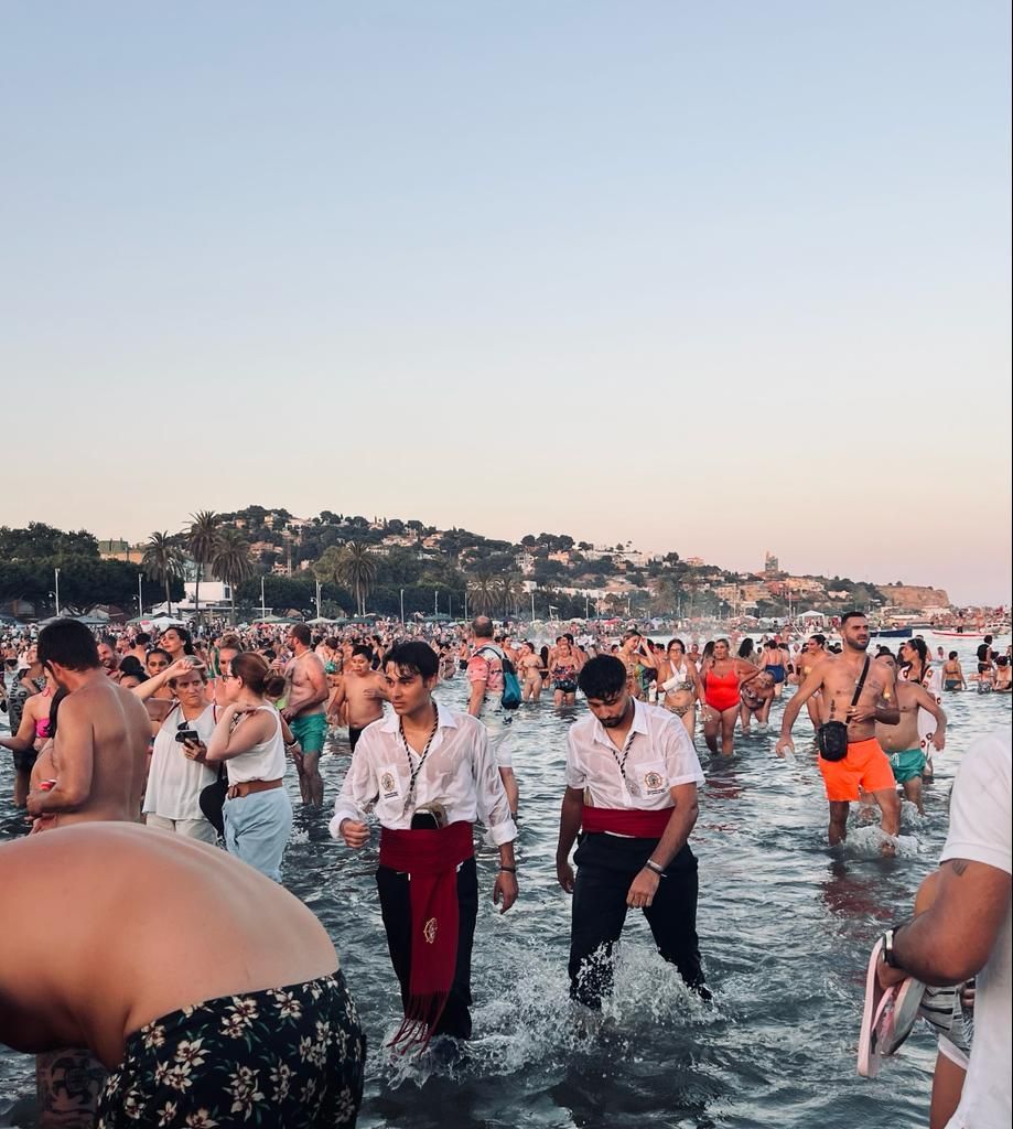 Personas chapoteando en las aguas poco profundas de la playa; muchos llevan bañadores y otras prendas ligeras.