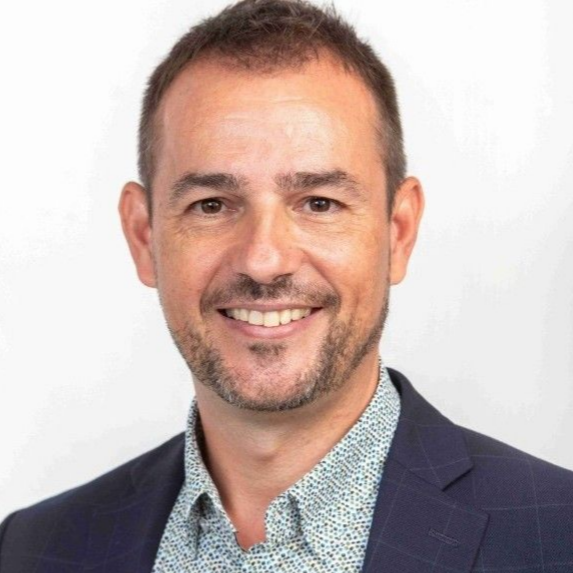 Hombre sonriente con blazer azul oscuro y camisa estampada, sobre fondo blanco.