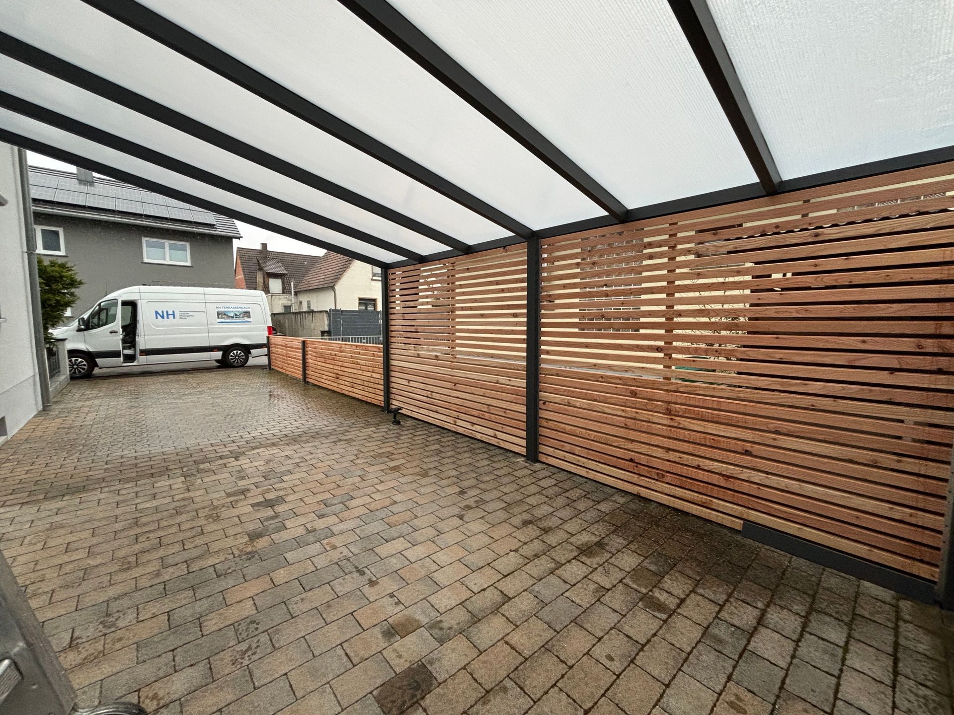Ein Carport mit Wänden aus Holzlatten, einem transparenten Dach und einem auf einer gepflasterten Auffahrt geparkten Lieferwagen.