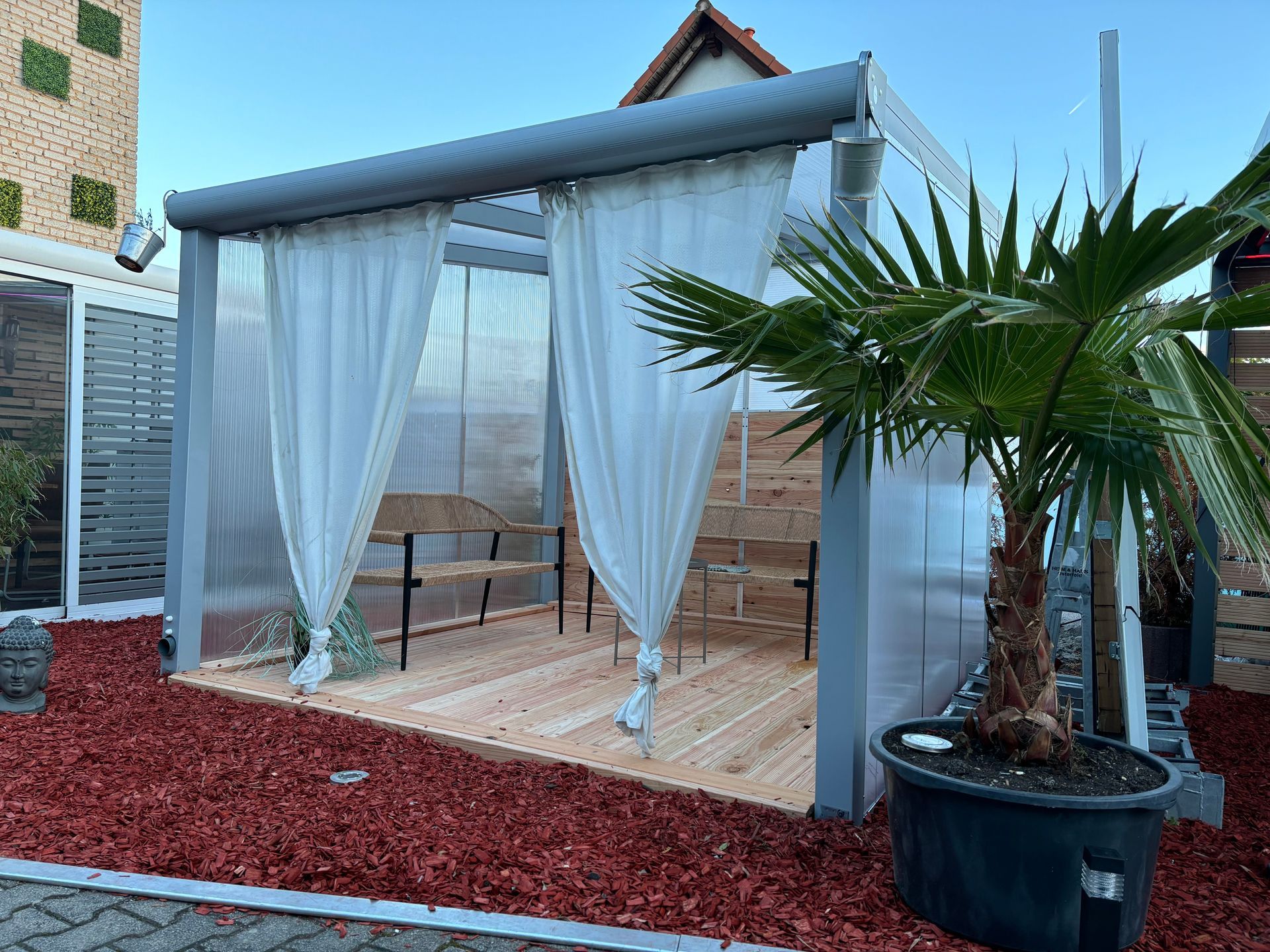 Ein Pavillon mit weißen Vorhängen, Holzboden und einer Palme. Roter Mulch umgibt das Bauwerk.