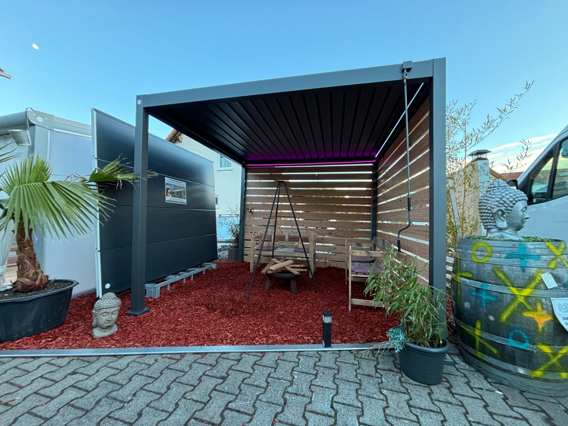 Ein Carport mit Wänden aus Holzlatten, einem transparenten Dach und einem auf einer gepflasterten Auffahrt geparkten Lieferwagen.