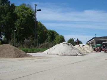 Baustelle mit Kieshaufen neben einer Straße, Bäumen und einem Traktor unter blauem Himmel.