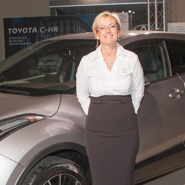 Une femme en chemise blanche et jupe noire se tient à côté d'une Toyota C-HR argentée.
