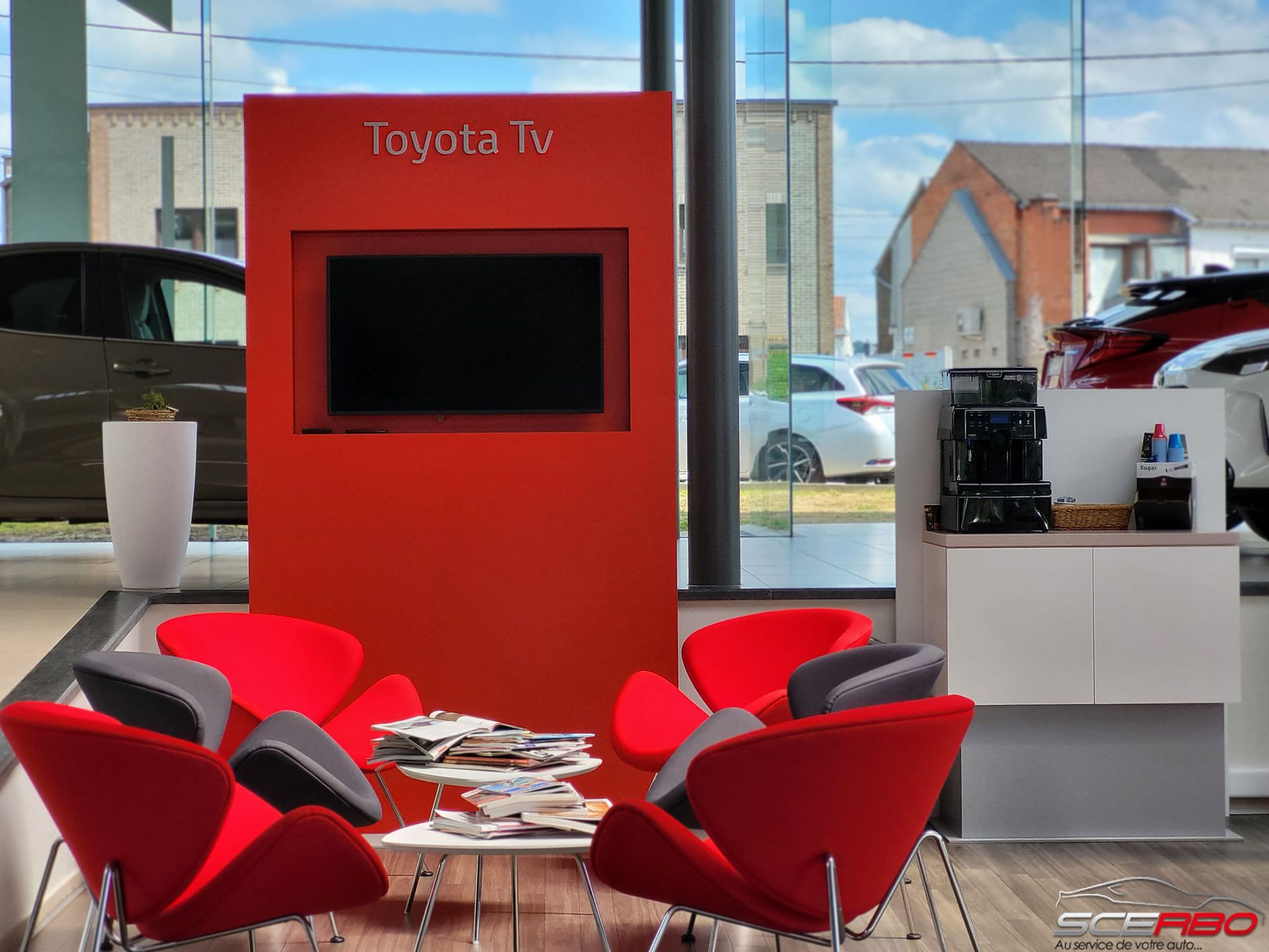Salle d'attente rouge dans une concession Toyota avec chaises, petite table, télévision et vue sur les voitures par la fenêtre.