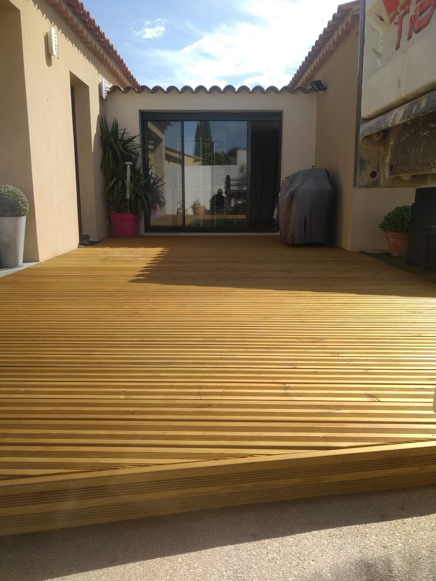 Une terrasse en bois mène à une maison aux portes vitrées. Journée ensoleillée, murs beiges, plantes en pot.